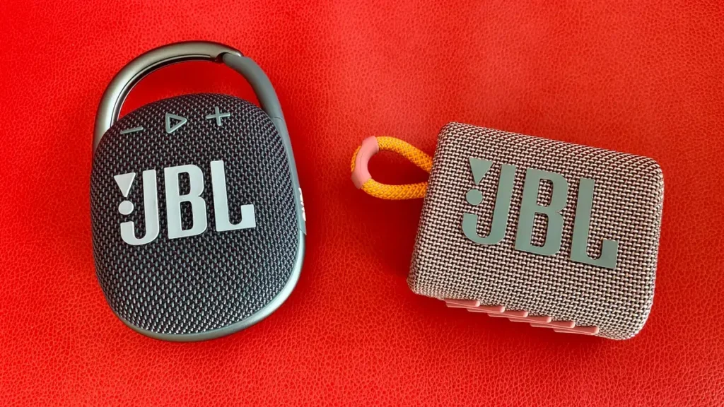 Black JBL Clip 4 and pink JBL Go 3 speakers on red background.